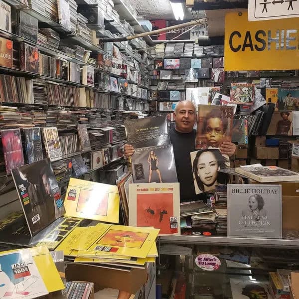 A record shop owner stands behind a crowded counter, holding up several vinyl records including albums by Mariah Carey, Sade, and Lauryn Hill.