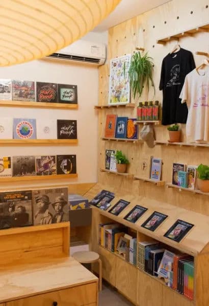 A modern record store corner with light wood shelving, vinyl records displayed on the wall, books and zines on angled racks, and potted plants adding greenery.