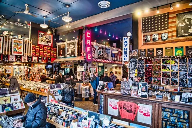 Busy, colorful record shop with a neon CAFE sign, posters and merch walls, and customers flipping through bins.