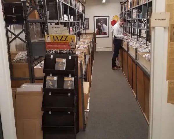 A man in a white shirt browses through tightly packed rows of jazz records inside a narrow shop lined with metal shelving.