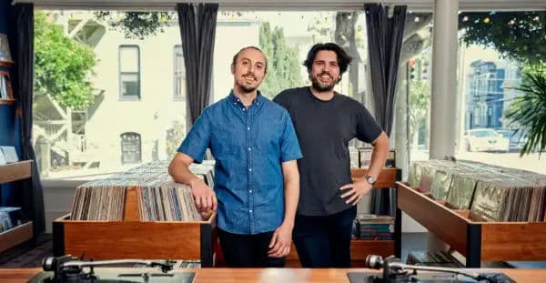 Two owners standing inside a bright Mission District record store with sunlight, large front windows, and rows of neatly organized vinyl bins.