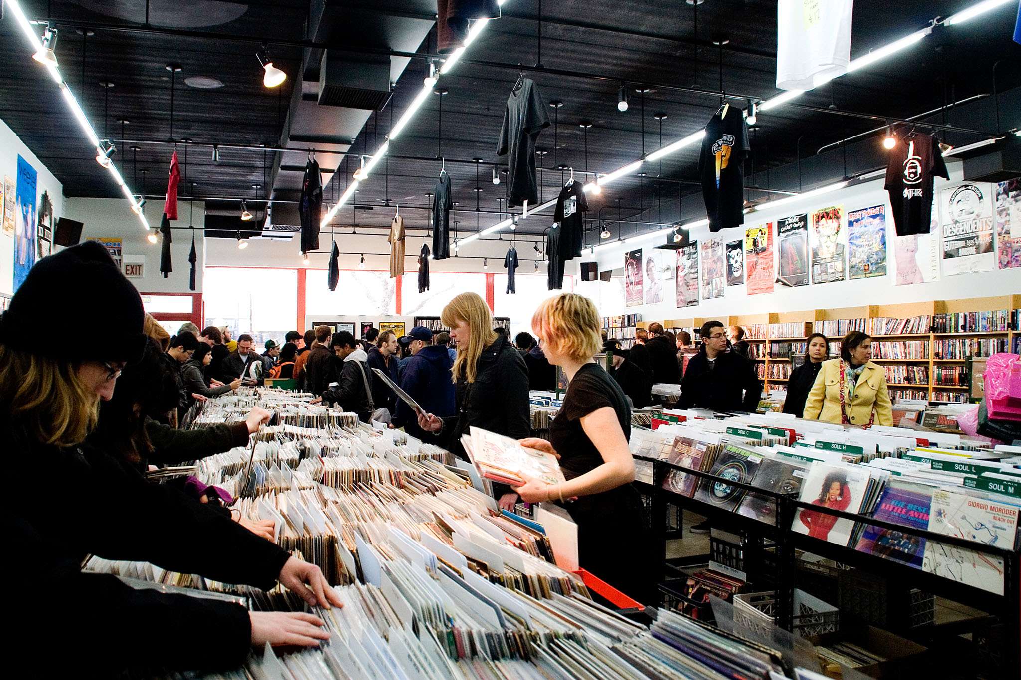 Crowded record store interior with shoppers flipping through vinyl bins at Reckless Records, with posters and hanging T-shirts overhead.