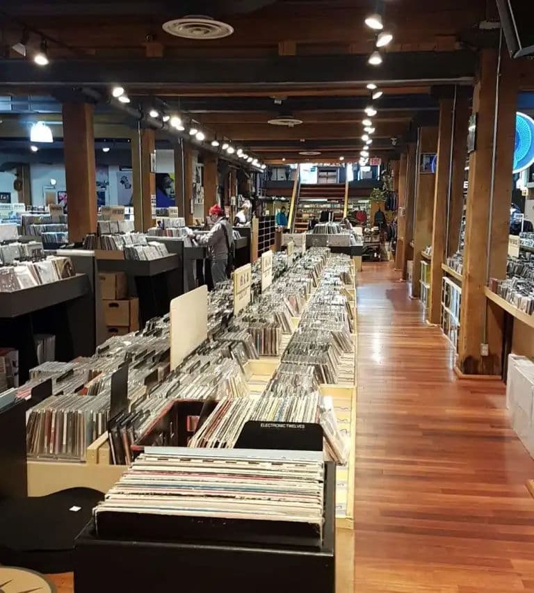 Long hardwood aisles lined with LP bins and CDs inside Sonic Boom, with shoppers browsing under exposed beams and track lights.