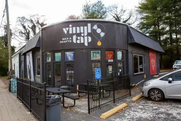 Exterior of Vinyl Tap, a record shop and bar with the tagline 'Wax & Drafts' painted on the building, with outdoor seating and a black facade.